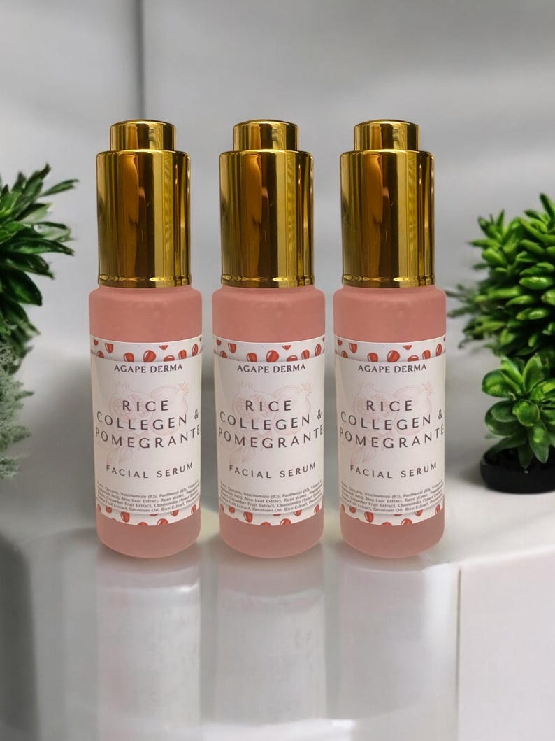 May include: Three pink facial serum bottles with gold-colored tops are displayed. Each bottle has a label with the text "RICE COLLAGEN & POMEGRANTE FACIAL SERUM". The bottles are arranged on a white surface with greenery in the background.