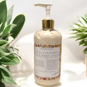Peut inclure: Une bouteille transparente de crème pour le corps au lait de chai et à la cannelle Agape Derma avec un distributeur à pompe blanc et des accents dorés. L'étiquette indique les ingrédients et la bouteille contient 355 ml de lotion. Des plantes vertes sont en arrière-plan.