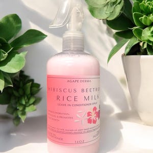 May include: A clear spray bottle with a pink liquid, labeled "Hibiscus Beetroot Rice Milk Leave In Conditioner Spray." The bottle has a white spray nozzle and a pink and white label with floral graphics. The bottle contains 14 oz of product.