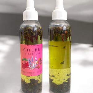 May include: Two clear glass bottles with white caps filled with a yellow liquid and herbs. The bottles are labeled "Agape Derma Chebe Hair Oil" with a pink background and a cherry, lavender, and flower design. The bottles are 4 oz.