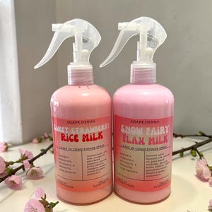 May include: Two pink spray bottles of hair conditioner. One is labelled "Sweet Strawberry Rice Milk" and the other "Snow Fairy Flax Milk." Both bottles have white spray nozzles and pink labels with product information. The bottles are on a white surface with pink flowers.