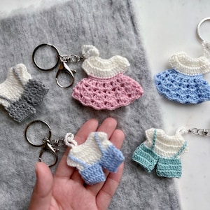 May include: Five crocheted keychains in the shape of dresses and overalls. The keychains are in various colors, including pink, blue, and green. Each keychain is attached to a silver key ring.