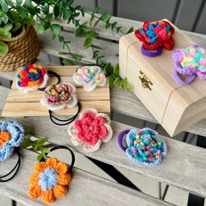 May include: Assortment of colorful crocheted flower hair scrunchies. The scrunchies are in various colors, including pink, blue, red, orange, and green. They are displayed on a wooden surface with a small wooden box.