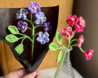 May include: Two bouquets of crocheted flowers in a vase and a black paper bag. The flowers are purple, pink, and green.