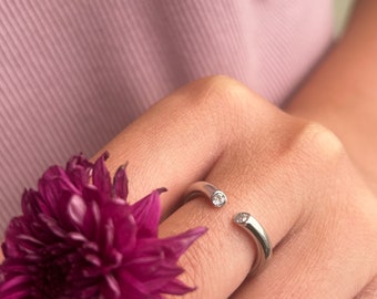Op de afbeelding: Een zilveren ring met twee kleine, heldere stenen aan de uiteinden. De ring is open en zit aan een vinger.