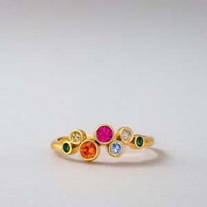 Könnte beinhalten: Ein goldener Ring mit einer Ansammlung farbiger Edelsteine. Der Ring hat ein lebendiges Design mit einem großen rosa Mittelstein, umgeben von orangefarbenen, blauen, grünen und weißen Edelsteinen. Der Ring ist vor einem schlichten weißen Hintergrund platziert.