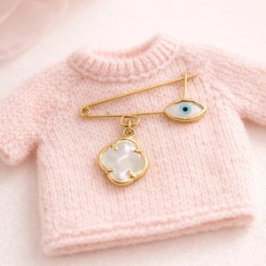 Könnte beinhalten: Ein kleiner, hellrosa Strickpullover mit einer goldfarbenen Sicherheitsnadel. Die Nadel hat einen weißen und goldenen vierblättrigen Kleeblatt-Anhänger und einen augenförmigen Anhänger mit einer blauen Iris. Der Pullover ist ein weicher, zarter Strick.