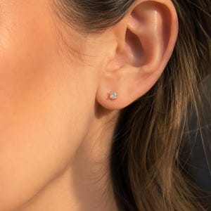 Pode incluir: Um pequeno brinco de pressão com uma pedra preciosa redonda e transparente, montado numa cravação de metal dourado e posicionado no centro do lóbulo da orelha. A pedra única facetada reflete a luz, criando um aspeto brilhante e cintilante.