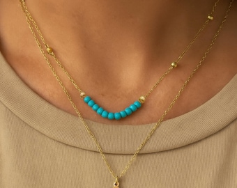 May include: A layered gold-tone necklace set featuring a short chain with small turquoise-coloured beads and a longer chain with a teardrop-shaped pendant containing a central turquoise-coloured stone and etched sunburst detailing.