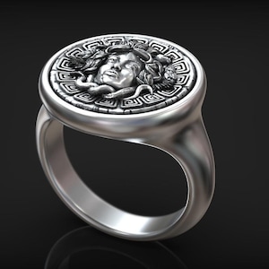 May include: A silver ring featuring a detailed relief of Medusa's face within a circular frame. The ring has a polished finish and a classic band design, suitable for everyday wear or special occasions.
