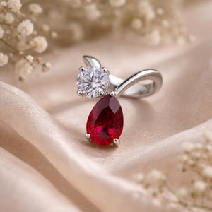 May include: A silver ring with a clear, round gemstone and a deep red, pear-shaped stone. The ring is set on a cream-coloured fabric, with small white flowers in the background.