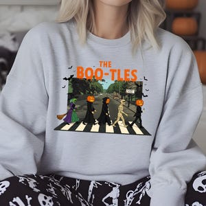 May include: Light gray sweatshirt with the text "The Boo-tles" in orange. The graphic depicts a Halloween-themed parody of the Beatles' Abbey Road album cover, with costumed figures crossing a crosswalk. Black bats are flying above.