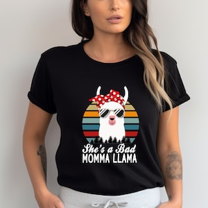 May include: Black t-shirt with a retro sunset graphic featuring a llama wearing sunglasses and a red bandana with white polka dots. The text "She's a Bad Momma Llama" is printed below the graphic.