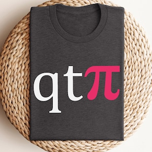 May include: Dark gray t-shirt with the text "qt π" in white and pink. The shirt is folded and placed on a round, woven surface. The logo "TEETOPIA UNIVERSE" is visible in the upper left corner.