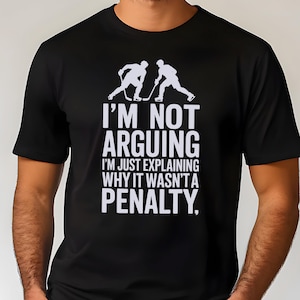 May include: Black t-shirt with white text and hockey player graphics. The text reads "I'M NOT ARGUING I'M JUST EXPLAINING WHY IT WASN'T A PENALTY." A fun, casual tee for hockey fans.
