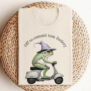May include: Beige t-shirt featuring a green frog wearing a purple wizard hat, riding a silver scooter. The text "Off to commit tom foolery" is arched above the frog. The shirt is displayed on a woven, circular surface.