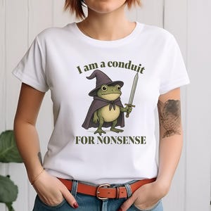 May include: White t-shirt featuring a cartoon frog wearing a wizard hat and cape, holding a sword. The text "I am a conduit FOR NONSENSE" is printed above and below the frog illustration. The shirt is a casual, everyday wear item.