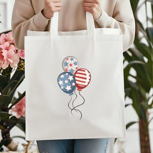 May include: White tote bag featuring a patriotic balloon design. The balloons are in red, white, and blue, with star and stripe patterns. The bag has long white straps and is held by a person.