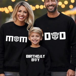 May include: A family matching set of black sweatshirts with white text and a robot graphic. The adult shirts say "MOM" and "DAD" and the child's shirt says "BIRTHDAY BOY".