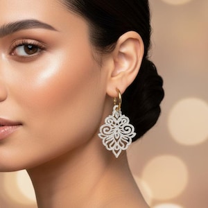 Könnte beinhalten: Eine Frau mit dunklem Haar, das zu einem Dutt gebunden ist, trägt verzierte, weiße und goldene Ohrringe. Die Ohrringe haben ein detailliertes, durchbrochenes Design. Der Hintergrund ist ein weiches, verschwommenes Bokeh aus warmen, goldenen Lichtern.