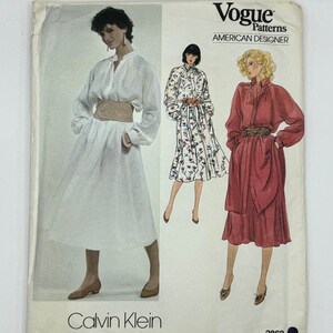 May include: Vintage Vogue Patterns sewing pattern featuring designs by Calvin Klein. The pattern includes illustrations of dresses with long sleeves and belted waists. The cover is off-white with the Vogue Patterns and American Designer logos.