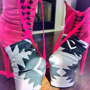 May include: A pair of high-heeled platform boots featuring a bright pink suede upper with black eyelets and neon pink laces. The lower portion of the boots is covered in a fabric with a black and white geometric zigzag pattern.