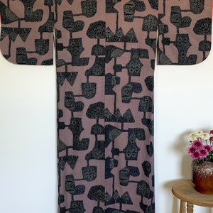 Pode incluir: Um quimono japonês tradicional com um padrão geométrico em preto e cinza sobre um fundo malva. O quimono tem mangas compridas e uma silhueta larga e fluida.