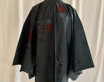 Op de afbeelding: Een donkergrijze jas in kimono-stijl met een bloemenpatroon. De jas heeft een textuurstof en is versierd met patches van bloemmotieven in rood en zwart. De mouwen zijn wijd en de jas wordt gepresenteerd op een houten mannequin.