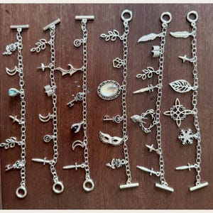 Puede incluir: Pulseras de plata con dijes variados, incluyendo lunas, llaves, hojas y dagas. Las pulseras se exhiben sobre una superficie de madera oscura, mostrando los intrincados detalles de cada dije.