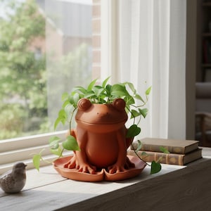 May include: A terracotta-colored ceramic planter shaped like a seated frog, resting on a matching lily pad-shaped base. The frog has prominent eyes and a wide mouth, with a green leafy plant growing out of the top of its head and cascading down.