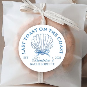 May include: White favour bags with a blue and white round sticker that says "Last Toast on the Coast" with a seashell design and the text "Est 2025 Beatrice's Bachelorette".