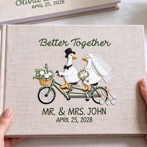 Könnte beinhalten: Ein beigefarbenes, leinengebundenes Hochzeitsgästebuch mit einer Stickerei von zwei Gänsen auf einem Tandemfahrrad. Der Text lautet Better Together, MR. & MRS. JOHN und APRIL 25, 2028. Die Gänse tragen Hochzeitskleidung, darunter einen Zylinder und Schleier.