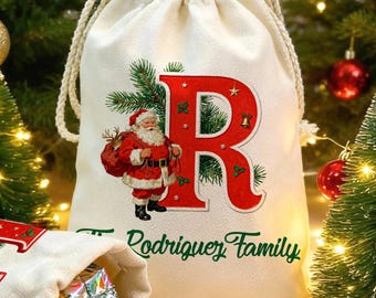 May include: Two off-white canvas drawstring gift bags featuring a large red letter R and E, each decorated with a graphic of Santa Claus, pine branches, and the text The Rodriguez Family, with small metallic gift boxes spilling out.