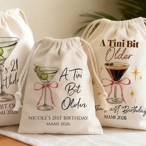 May include: Three off-white drawstring bags with cocktail designs. One features a martini with olives and the text "Martin's 21st Birthday." Another says "A Tini Bit Older" with a martini and a pink bow. The third bag has a similar design with the text "Nicole's 21st Birthday."