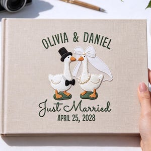 Könnte beinhalten: Ein beiges, stoffbezogenes Fotoalbum mit einer Stickerei von zwei Gänsen, die für eine Hochzeit gekleidet sind. Der Text lautet OLIVIA & DANIEL, Just Married und APRIL 25, 2028. Die Gänse tragen einen schwarzen Zylinder, eine Fliege, einen Schleier und eine Perlenkette.