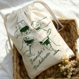 May include: Cream-coloured drawstring bag featuring a green illustration of hands holding cocktail glasses. The text "Jennifer's Bachelorette" is printed in green script. The bag is in a woven basket.