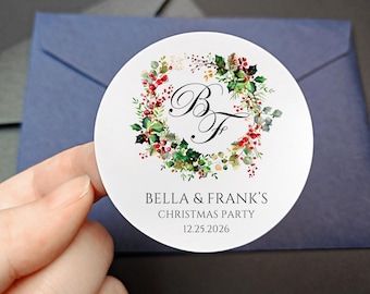 May include: A round white sticker with a watercolor wreath of green leaves and red berries. The sticker has the initials "B F" in elegant script, with the text "BELLA & FRANK'S CHRISTMAS PARTY 12.25.2026". The sticker is held over a blue envelope.