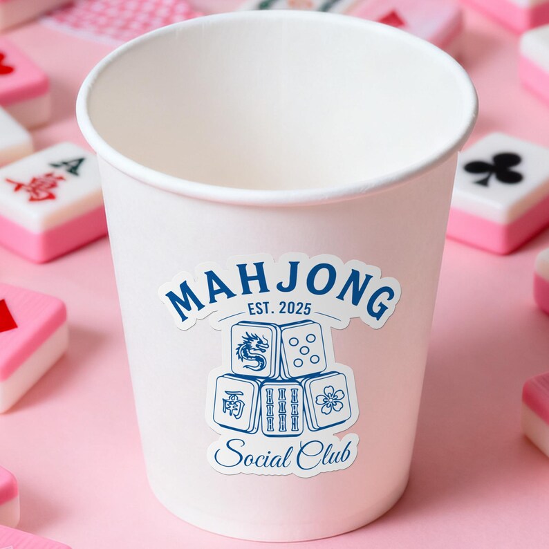 Może przedstawiać: Biały papierowy kubek z niebiesko-białą naklejką "Mahjong Social Club". Naklejka zawiera ilustracje kości i tekst "Est. 2025". Kubek otoczony jest r&oacute;żowymi i białymi kafelkami do gry w Mahjonga.