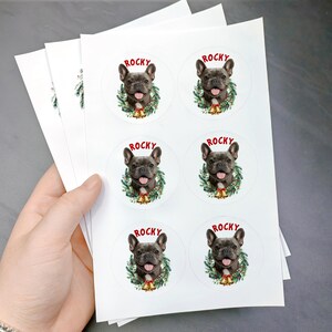 Op de afbeelding: Vellen witte stickers met een Franse Bulldog met de naam "Rocky" in het rood. Elke sticker toont de hond met een groene krans en een gouden bel. De stickers zijn rond en worden door een hand vastgehouden.