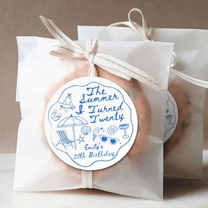 Könnte beinhalten: Zwei weiße Papiertüten, gebunden mit cremefarbenem Band, die jeweils eine Leckerei enthalten. Ein blau-weißer runder Aufkleber auf der Vorderseite jeder Tüte lautet "The Summer I Turned Twenty" mit Strandmotiven und "Emily's 20th Birthday".