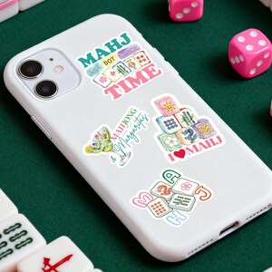 Może przedstawiać: Biały smartfon z wieloma kolorowymi naklejkami. Naklejki zawierają tekst i obrazy związane z Mahjongiem, takie jak kostki, kafelki i koktajle. R&oacute;żowe kostki do gry są rozrzucone wok&oacute;ł telefonu na zielonej powierzchni.