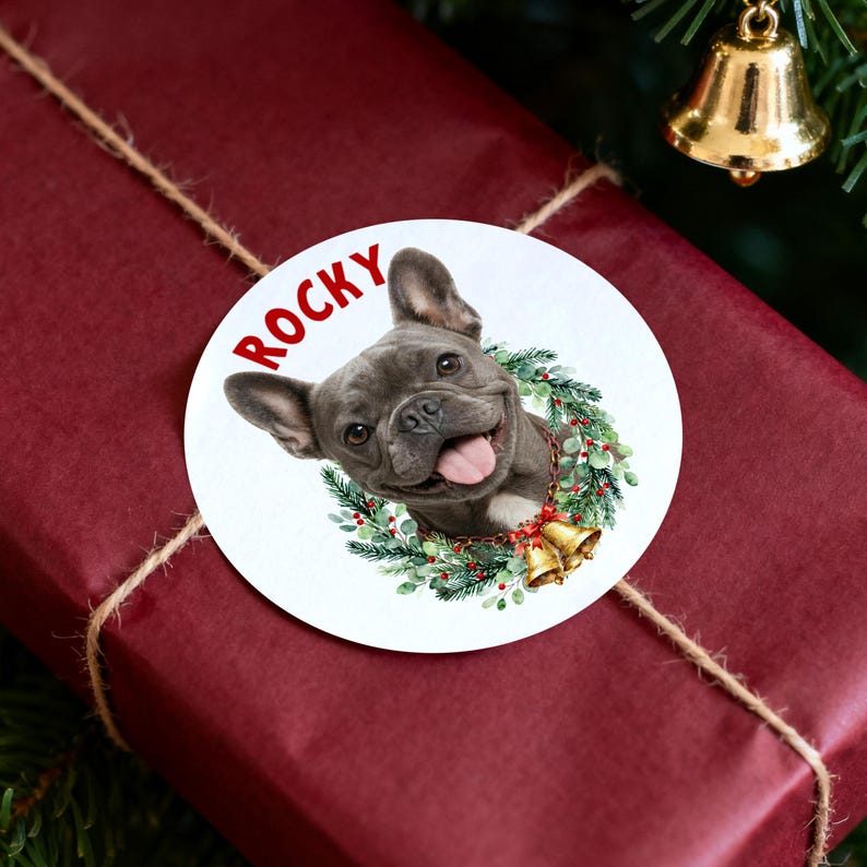 Op de afbeelding: Een ronde sticker met de naam "Rocky" in rode tekst. De sticker toont een grijze Franse Bulldog met een kerstkrans en belletjes. De sticker zit op een rood ingepakt cadeau.