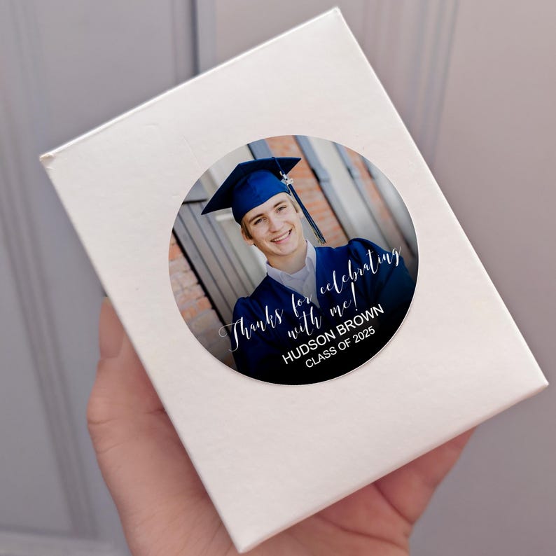 Peut inclure: Autocollant rond avec une photo d'un dipl&ocirc;m&eacute; portant une toque et une robe bleues. Le texte sur l'autocollant est "Merci de c&eacute;l&eacute;brer avec moi ! HUDSON BROWN CLASSE DE 2025".