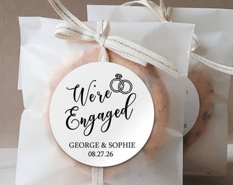 May include: White favor bags with a round sticker that says "We're Engaged" with a ring design and the names George & Sophie and the date 08.27.26.