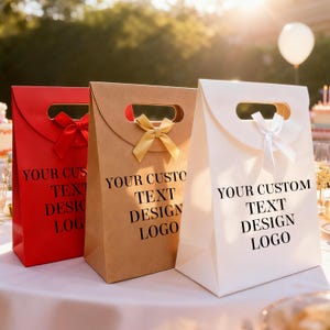 May include: Three gift bags in red, tan, and white, each with a handle and a bow. The bags have the text "YOUR CUSTOM TEXT DESIGN LOGO" printed in black. The bags are on a white tablecloth.