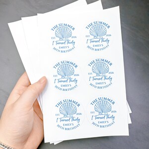 May include: A stack of white stickers with a blue seashell design and text. The text reads "THE SUMMER EST. 2026 I Turned Thirty EMILY'S 30TH BIRTHDAY". The stickers are round and are held in a person's hand.