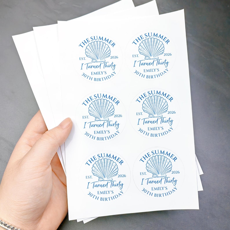May include: A stack of white stickers with a blue seashell design and text. The text reads "THE SUMMER EST. 2026 I Turned Thirty EMILY'S 30TH BIRTHDAY". The stickers are round and are held in a person's hand.