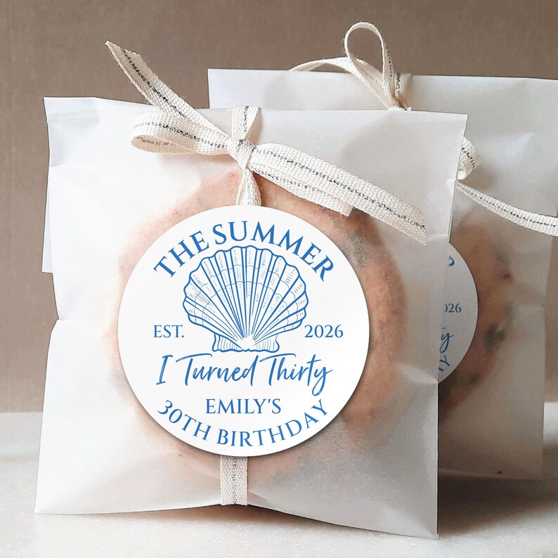 May include: Two white paper bags tied with cream-colored ribbon, each holding a cookie. A round blue and white sticker on the front of each bag reads "The Summer Est. 2026 I Turned Thirty Emily's 30th Birthday" with a seashell graphic.