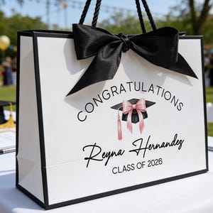 May include: White graduation gift bag with black trim, handles, and a large bow. The bag features the text "Congratulations Reyna Hernandez CLASS OF 2026" and a graduation cap graphic with a pink bow and tassel.