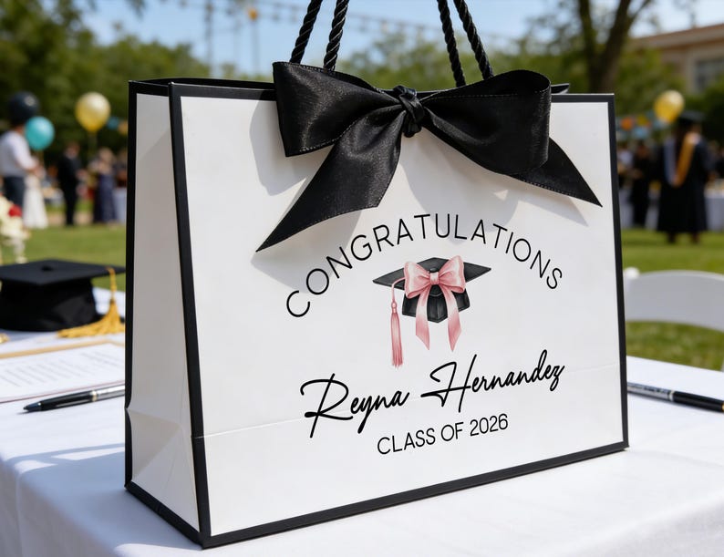 May include: White graduation gift bag with black trim, handles, and a large bow. The bag features the text "Congratulations Reyna Hernandez CLASS OF 2026" and a graduation cap graphic with a pink bow and tassel.